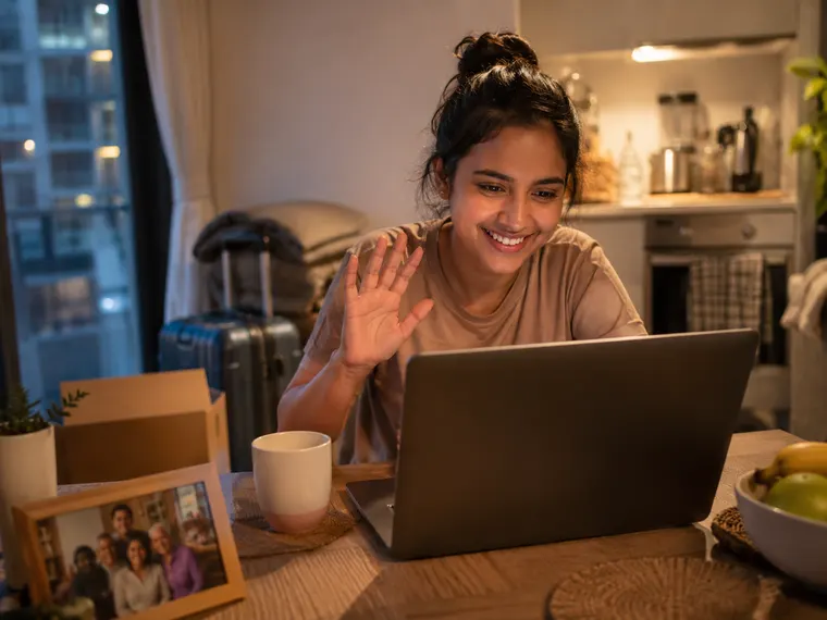 A young woman on a video call at her laptop at home, a packed suitcase behind her
